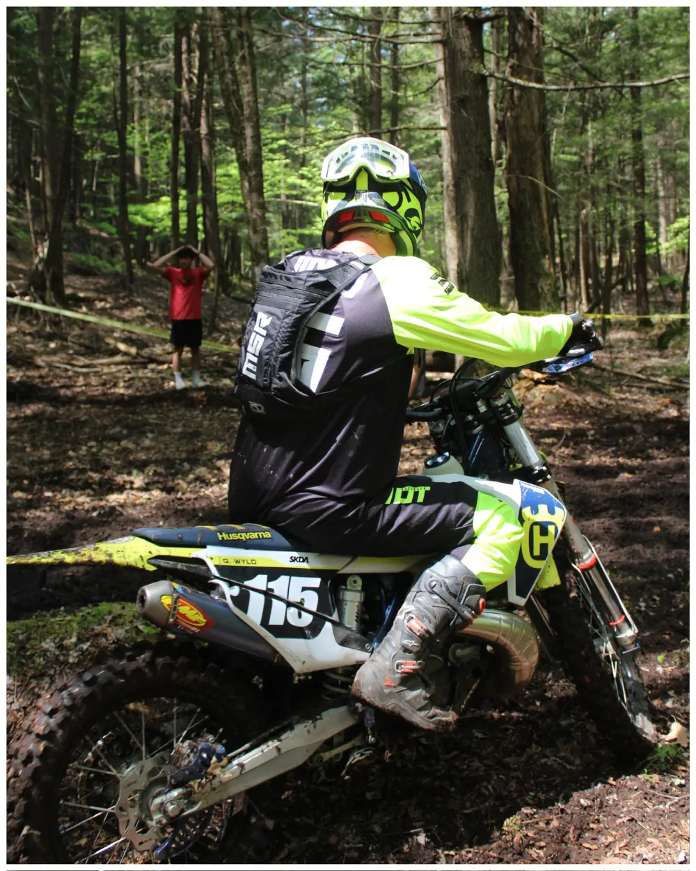 Motocross rider numbered 115 riding along a wooded trail during an event